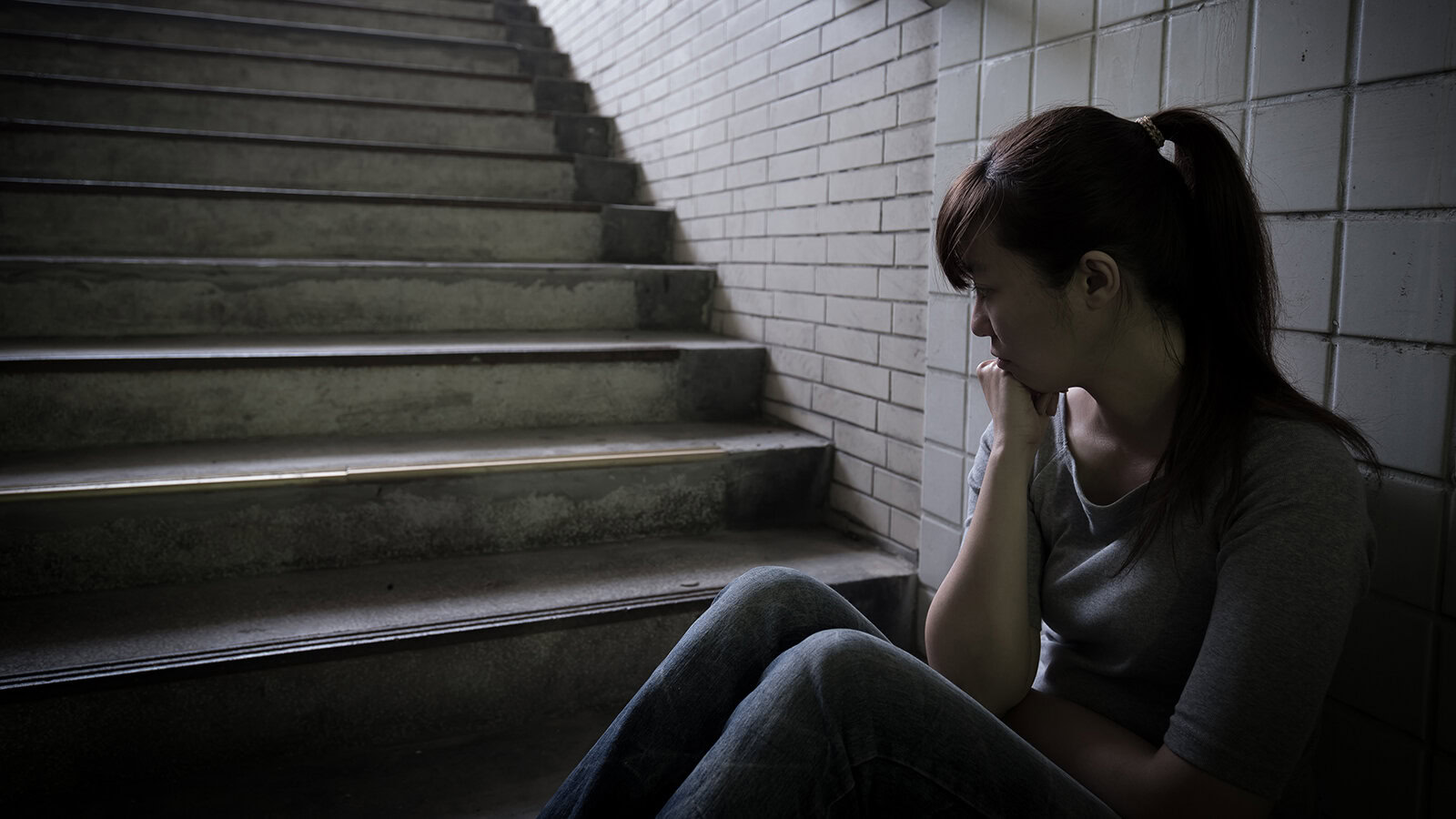 Feeling distressed and lonely, a woman sits against a brick wall near concrete stairs in an urban setting, illustrating emotional struggles and mental health awareness promoted by the Singapore Kindness Movement.