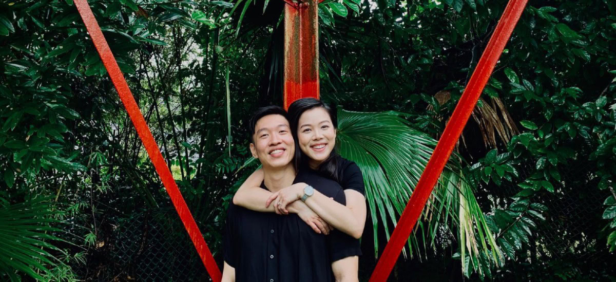 Brilliant couple embracing in lush green garden with red metal structure promoting Singapore Kindness Movement.