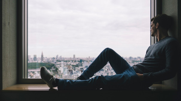 man sitting on brown wood windowsill looking out to buildings across