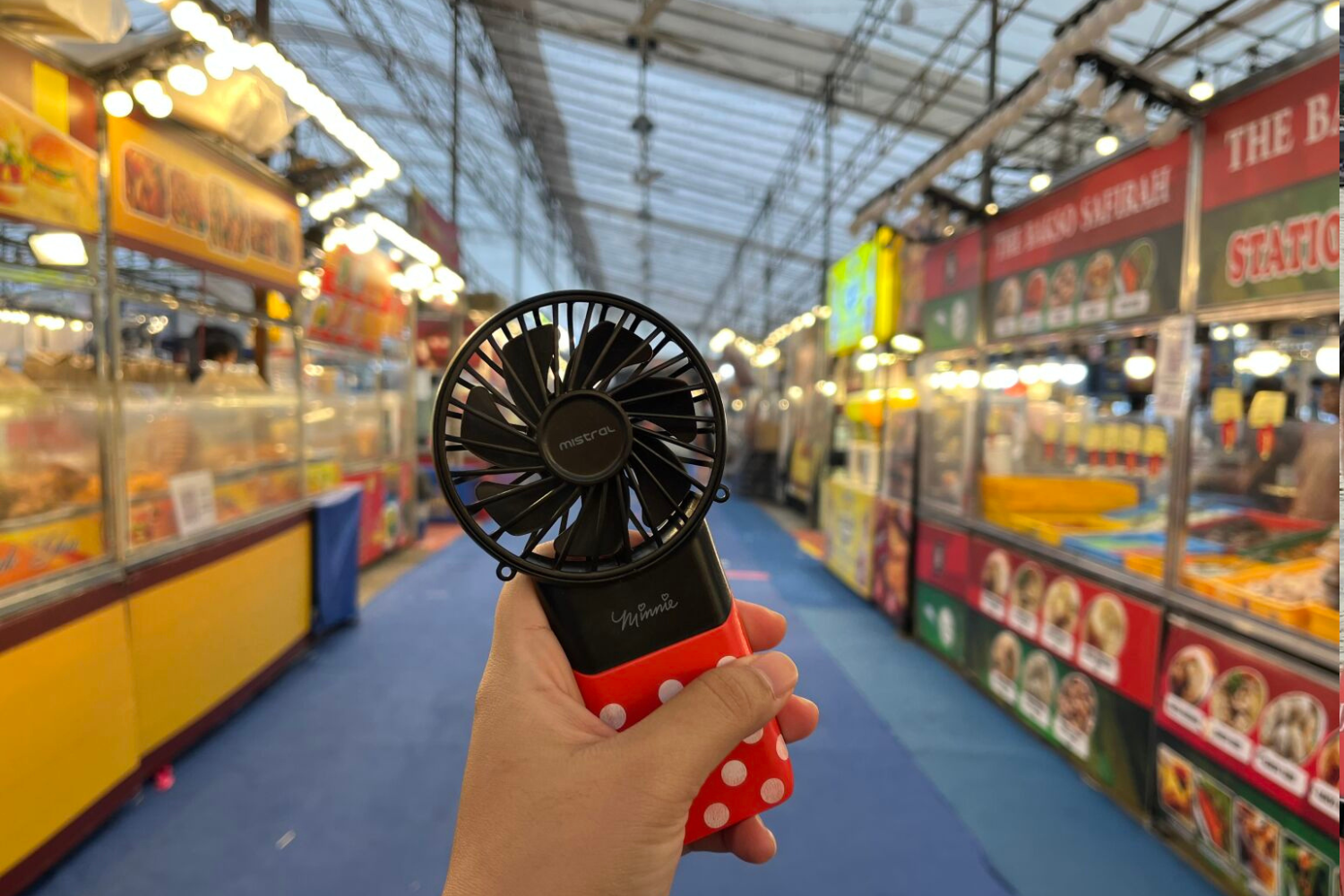 Mini handheld fan being held by a person inside a busy market or food stall area in Singapore. The fan is black with a red polka dot handle and branded "Mistral" and "Yummee". The background shows colorful stalls, illuminated signs, and a high, glass-paneled ceiling, capturing the lively atmosphere of Singapore markets, emphasizing community, kindness, and vibrant local culture.
