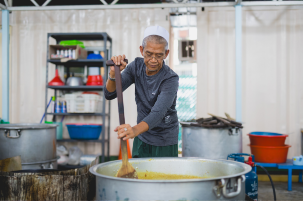 Man in a songkok cooking briyani in a big pot for an iftar event.