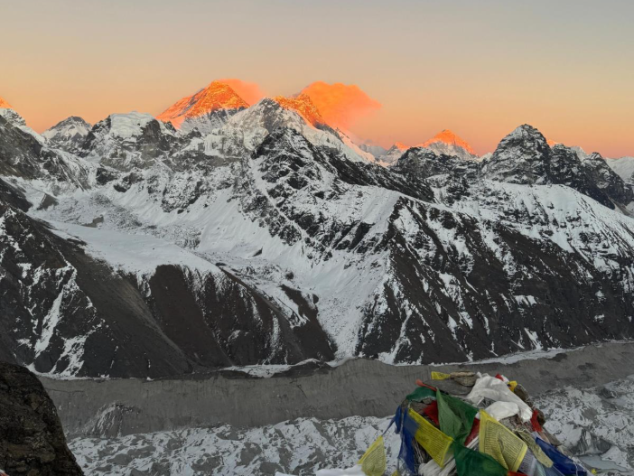 Climber resting on snowy mountain peak with colorful prayer flags, overlooking a stunning range of snow-capped mountains at sunrise, symbolizing resilience, hope, and unity in challenging environments.