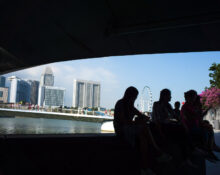People sitting on a bench under a bridge with Singapore skyline and river in the background, celebrating Pride and kindness in Singapore.