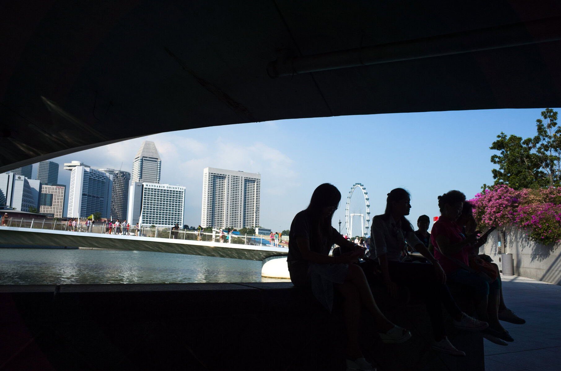 People sitting on a bench under a bridge with Singapore skyline and river in the background, celebrating Pride and kindness in Singapore.
