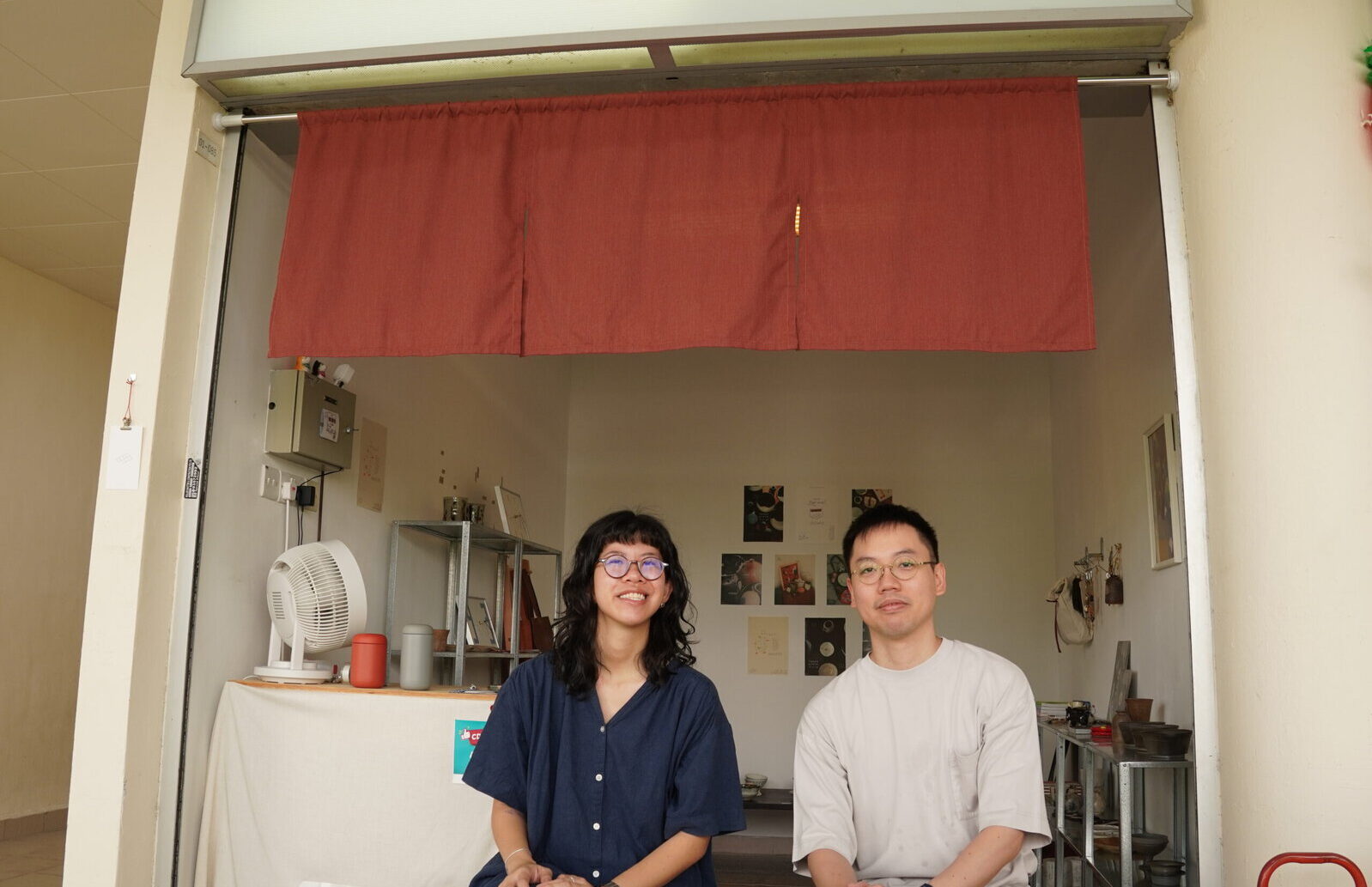 Two young adults standing inside a community store, smiling at the camera, with shelves and items in the background.