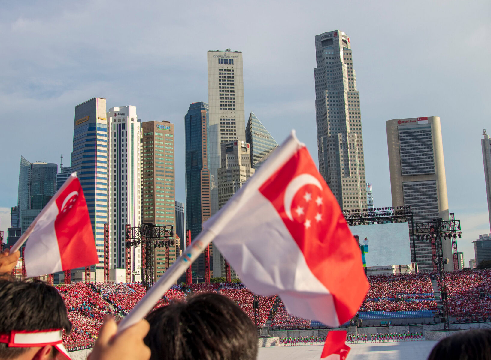 Crowd waving Singapore flags during National Day celebration in front of Singapore's skyline.