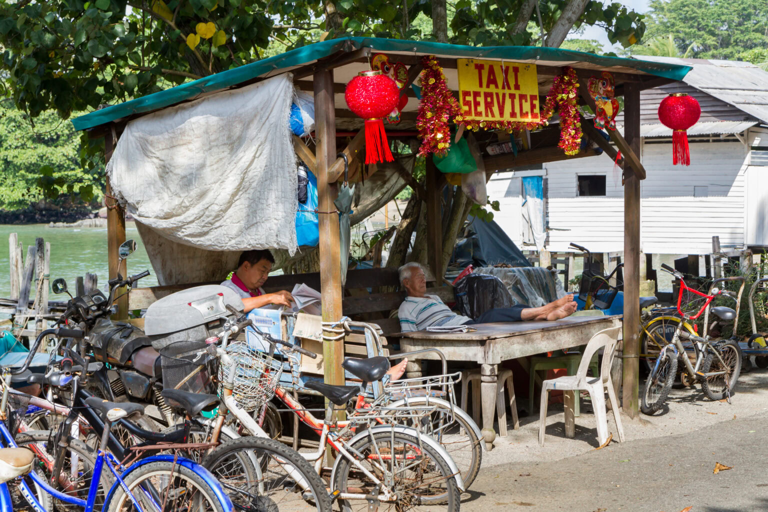 A small roadside taxi service stand with bicycles parked in front, two elderly men resting under a makeshift shelter, surrounded by greenery and simple houses.