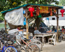 A small roadside taxi service stand with bicycles parked in front, two elderly men resting under a makeshift shelter, surrounded by greenery and simple houses.